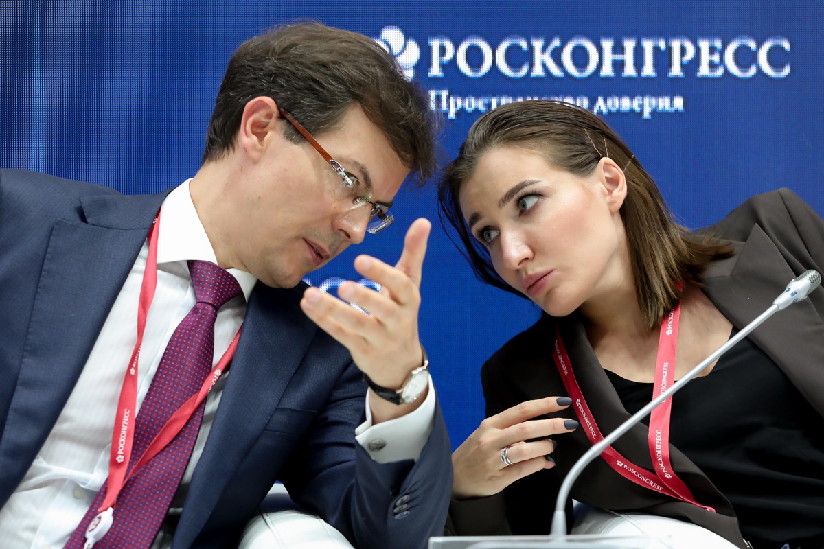 Два делегата форума разговаривают, наклонившись друг к другу рядом с конференционными микрофонами.