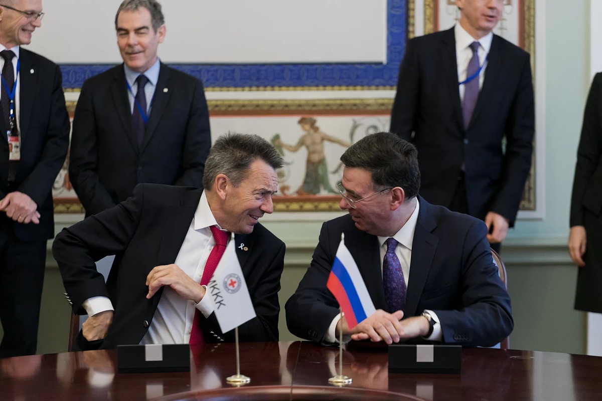 Делегаты разговаривают за формальным столом переговоров, перед ними флаги.