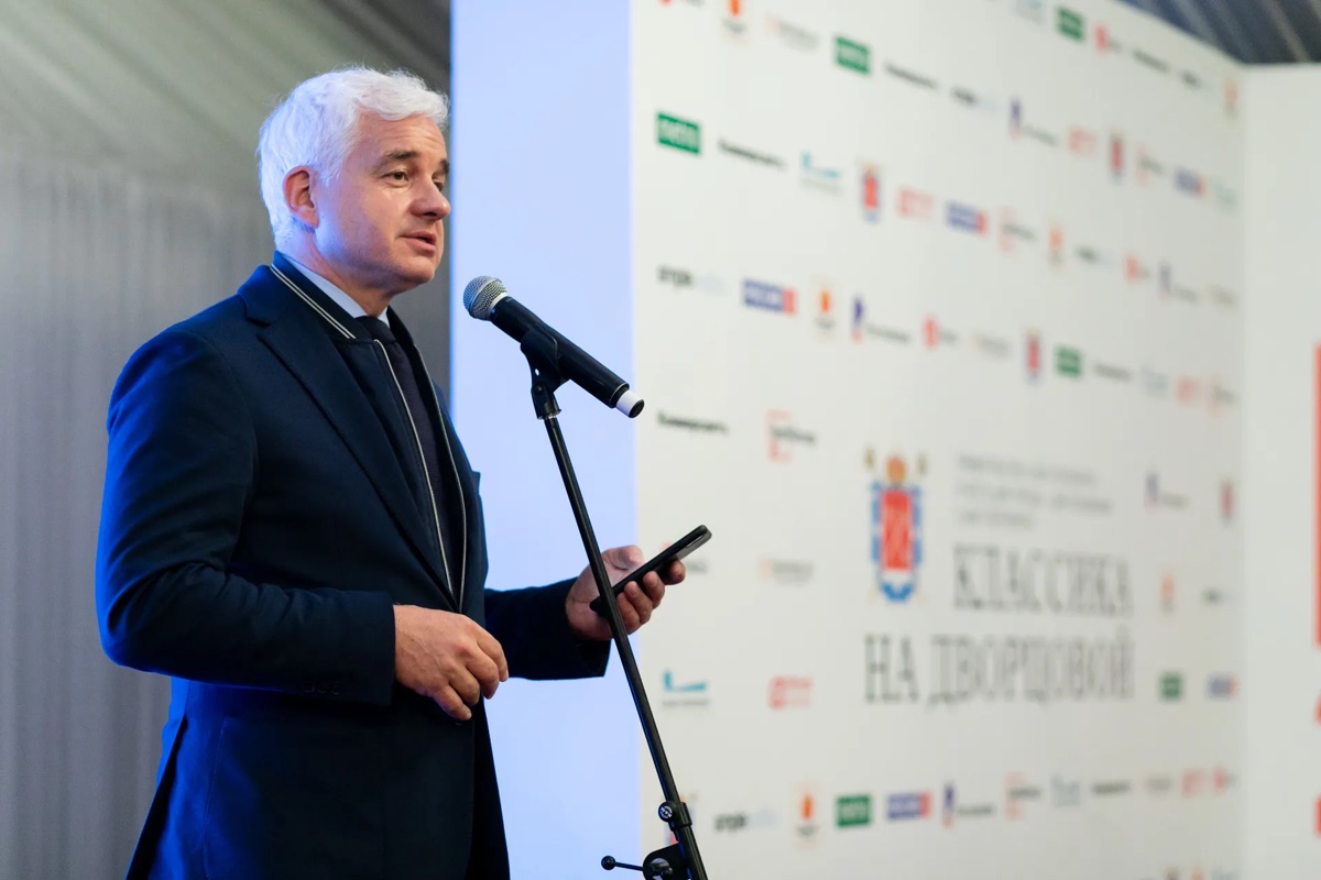 Спикер стоит у микрофона перед брендированным фоном мероприятия.