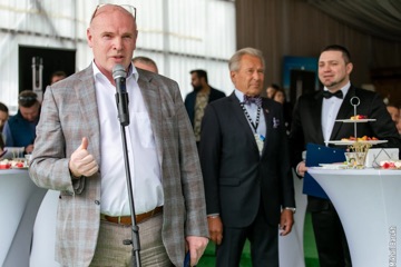 Спикер держит микрофон во время приема на открытом воздухе, гости стоят позади.