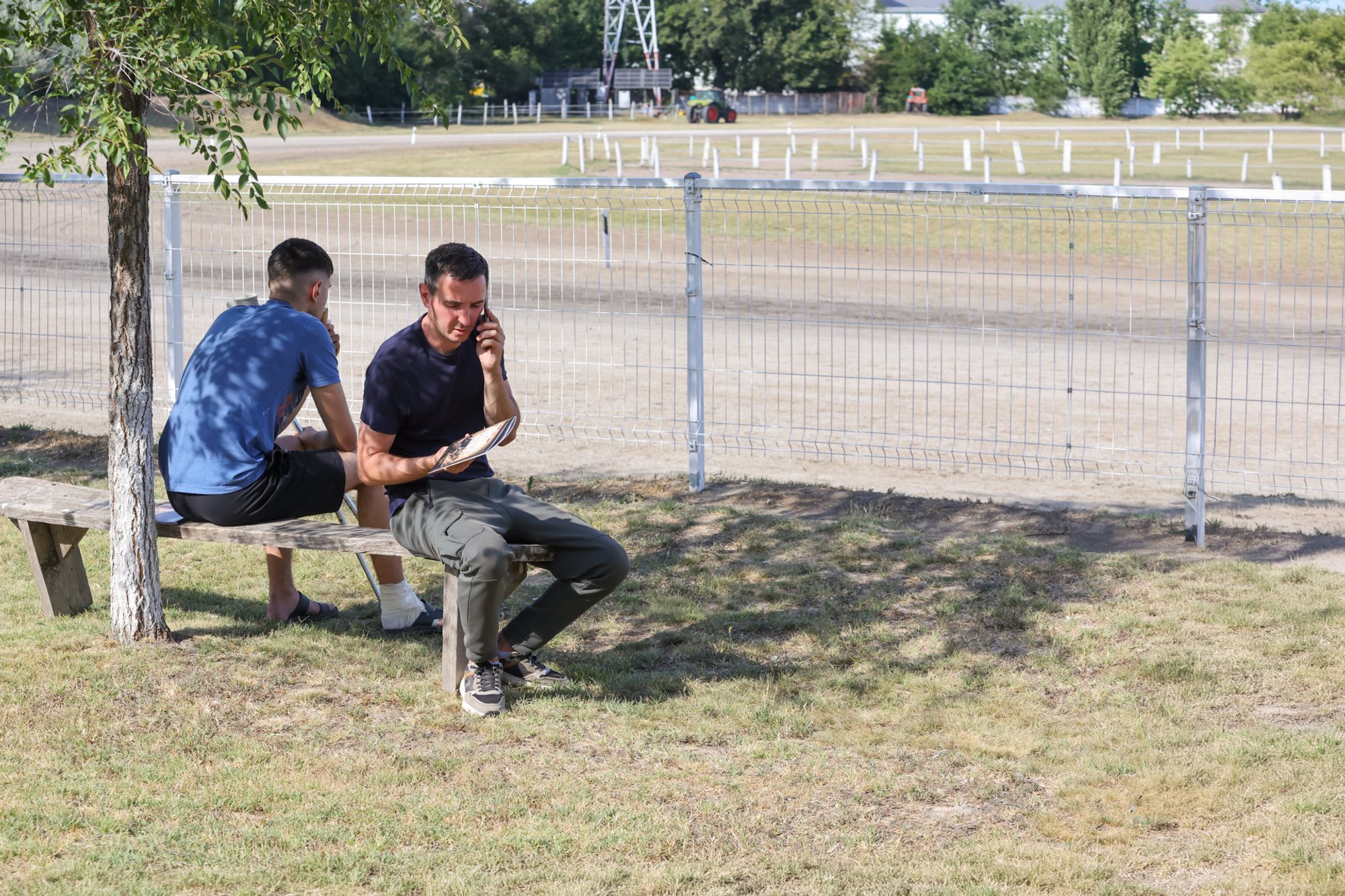 Two people seated on a wooden bench under a tree beside a fenced trotting track; white railings and empty starting gates visible across the track.