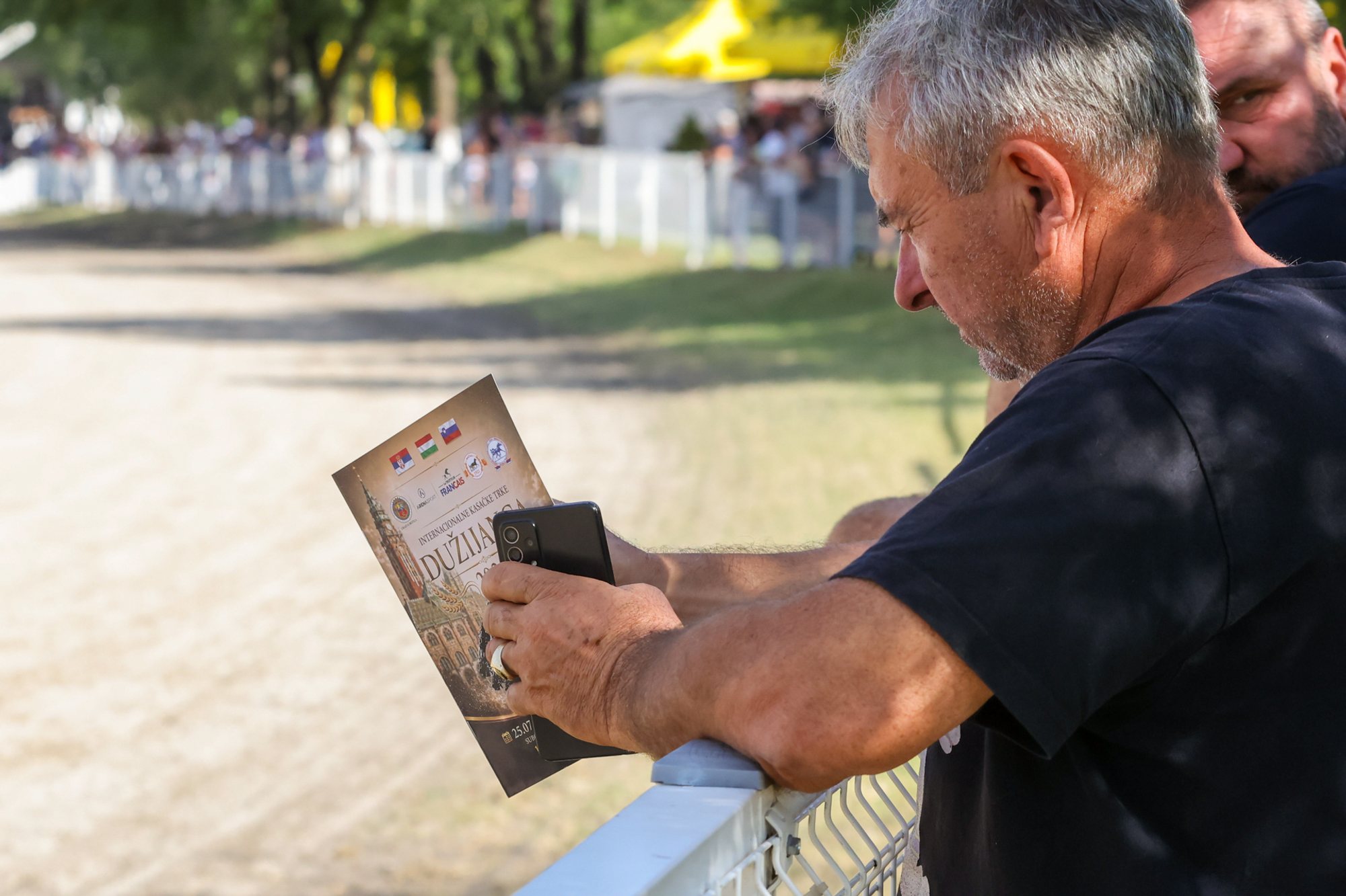 Two people leaning on a metal railing beside an unpaved racetrack; one holds a folded event programme and a smartphone, with stands and yellow tents blurred in the background.