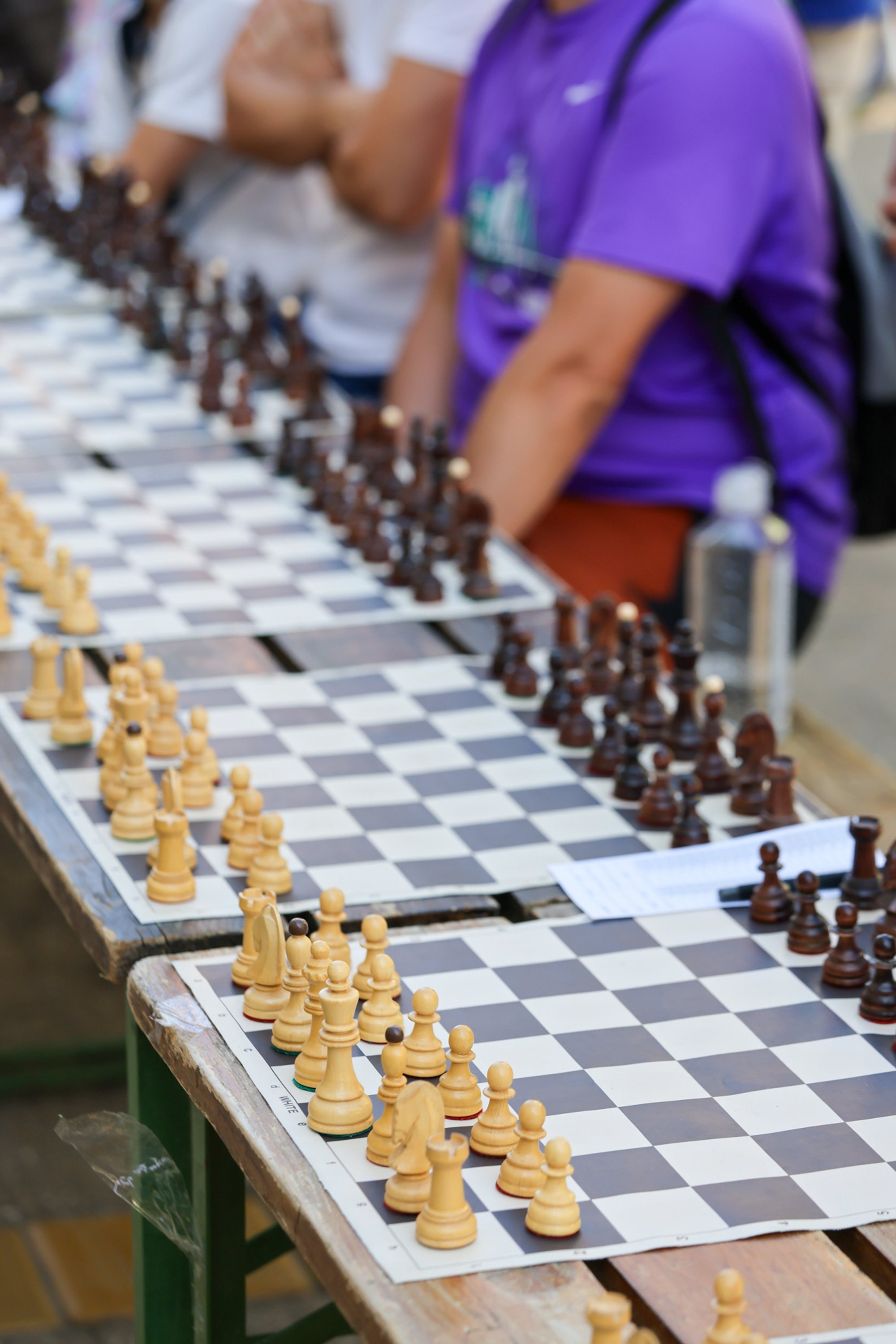 Multiple chessboards lined on folding tables with white and black pieces set for play; seated people are visible out of focus along the row.