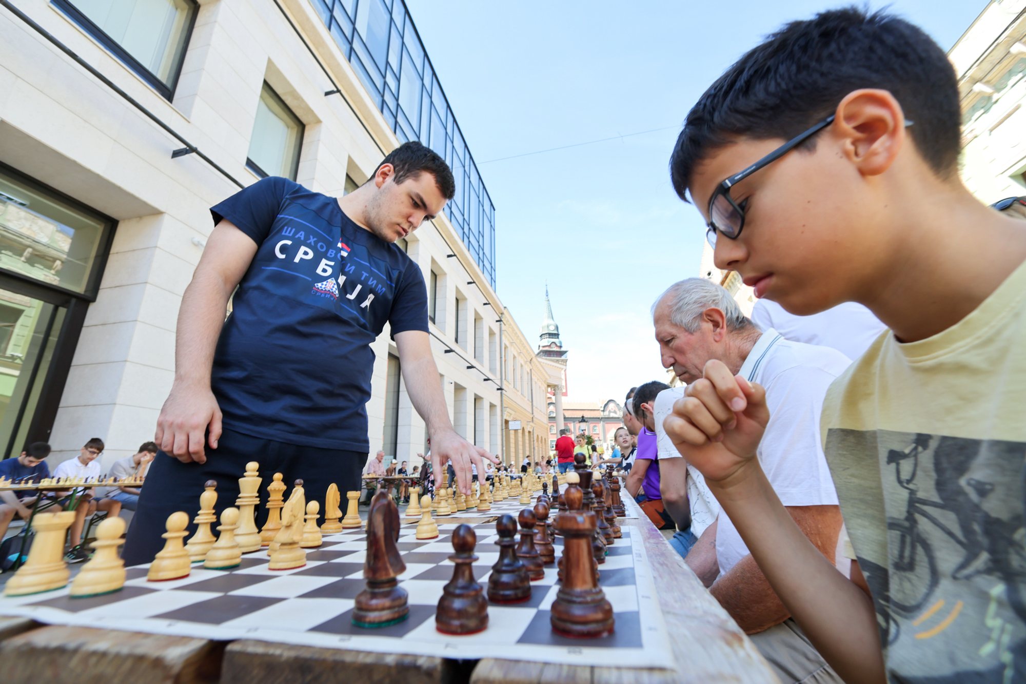 Outdoor row of chessboards on a long table; a boy with glasses looks at a board while an adult stands opposite; pedestrians and buildings in the background.