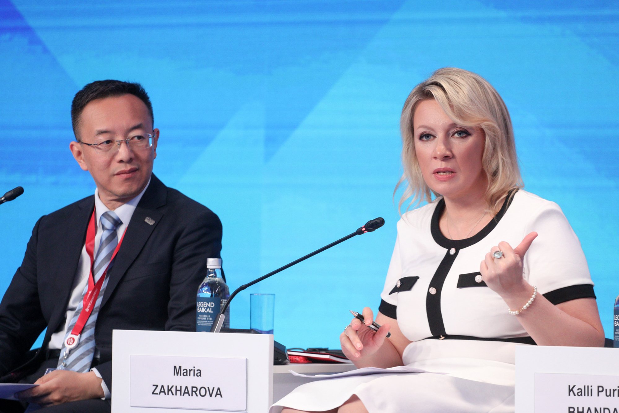 Two panel participants seated at a conference table against a blue stage backdrop; one person speaks into a tabletop microphone while the other sits beside them, with a water bottle and a visible nameplate on the table.