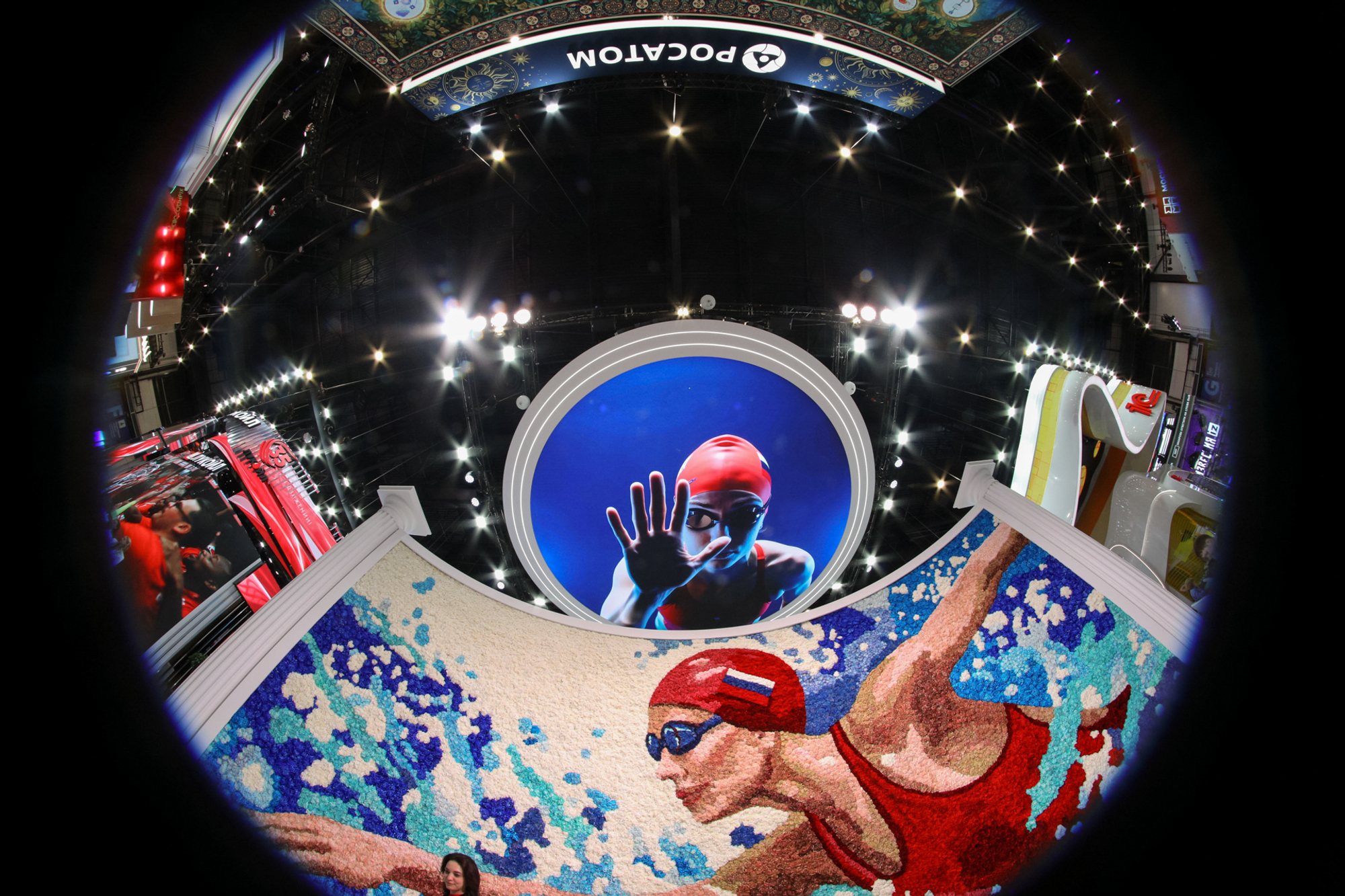 Low-angle view inside an ExpoForum exhibition booth: a curved foreground panel printed with stylized swimmers in red swim caps and, above it, a large circular overhead screen showing a person in a red helmet; ceiling spotlights and adjacent booth structures surround the display.