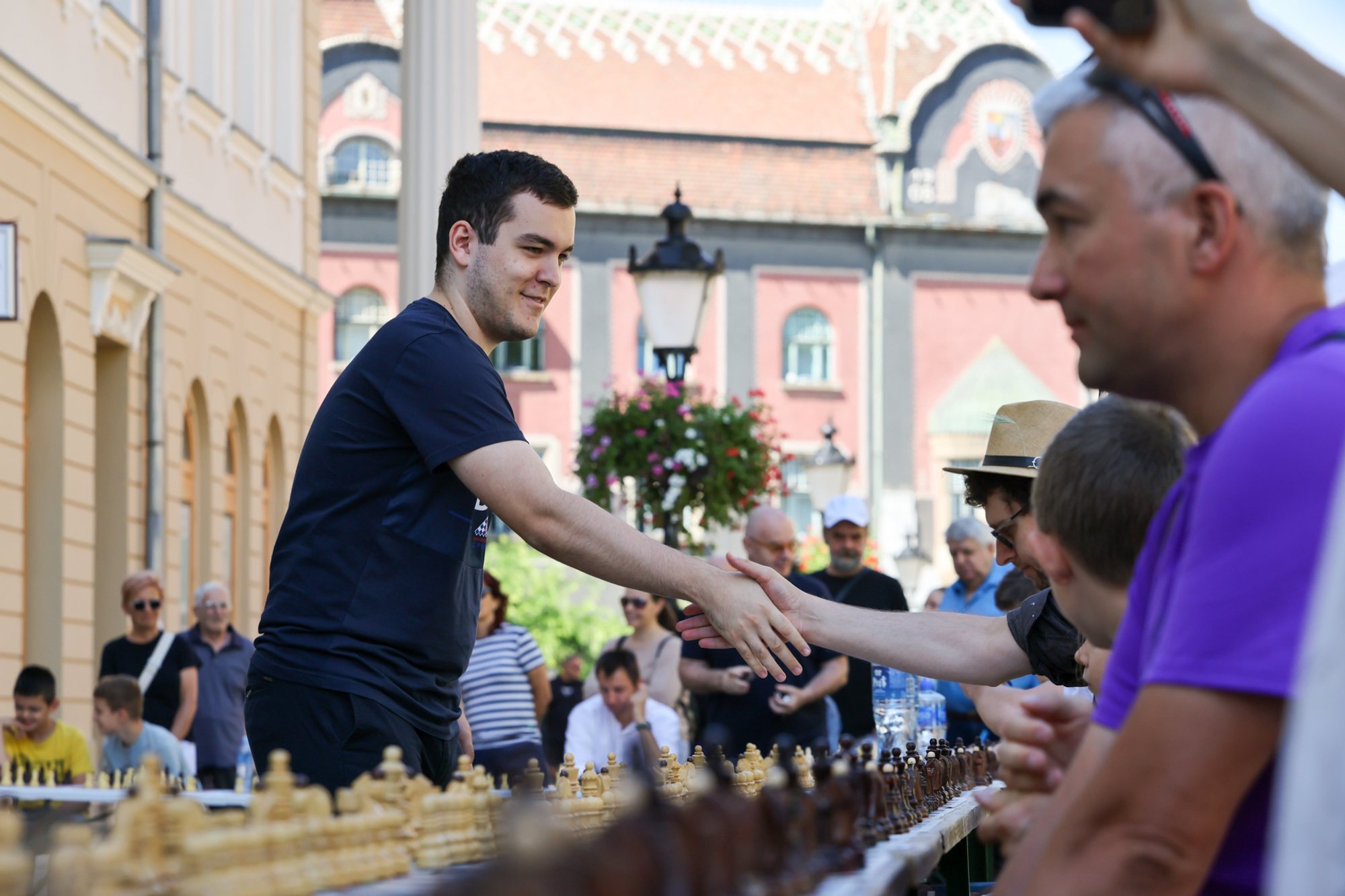 A man reaches along a long outdoor row of chessboards to shake hands with another person at Subotički Korzo; tables with chess pieces, a standing crowd and historic façades are visible.
