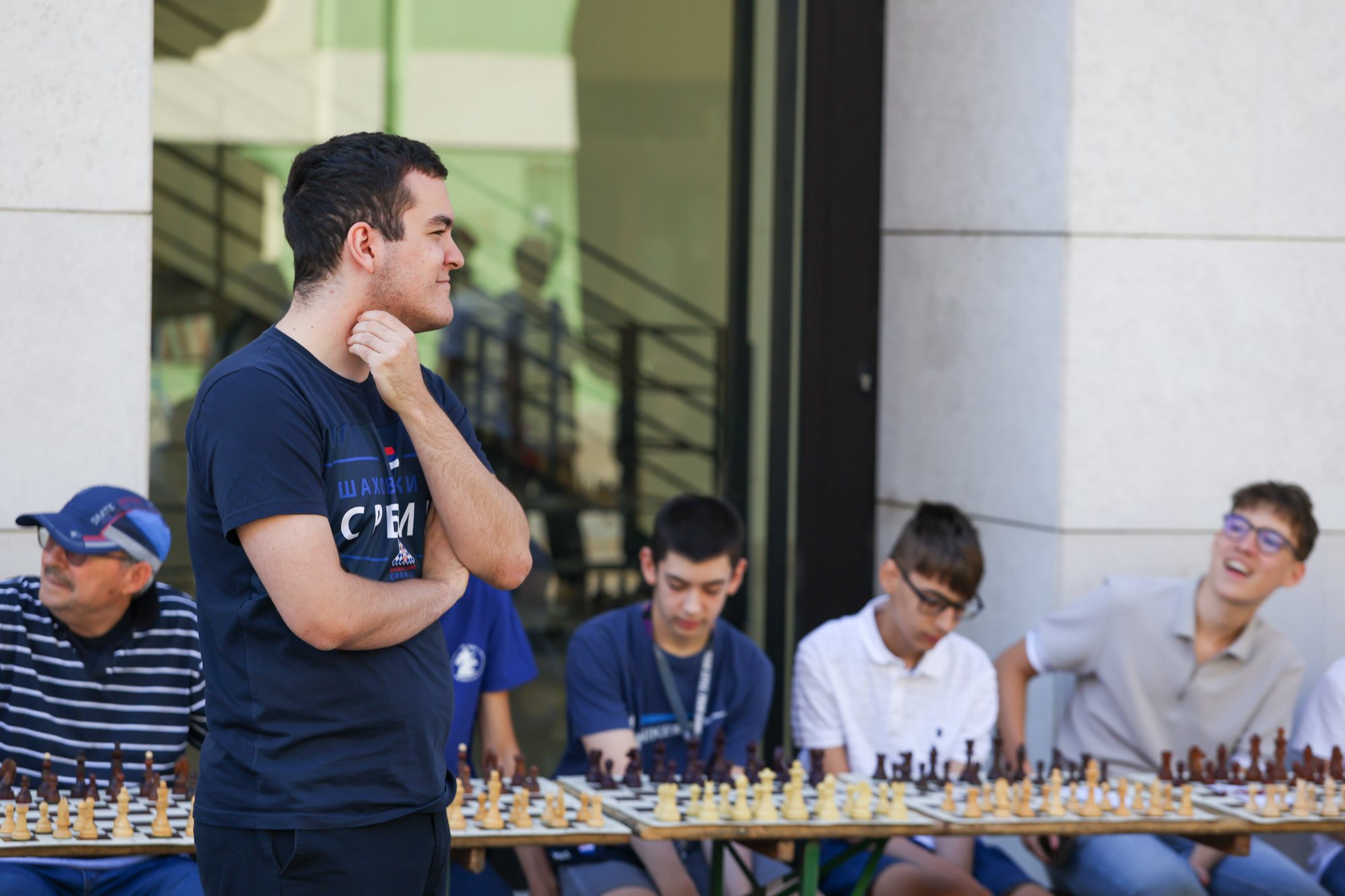 Man standing with his hand on his neck beside a long row of chessboards on an outdoor table; four seated players facing the boards and a building entrance behind them.