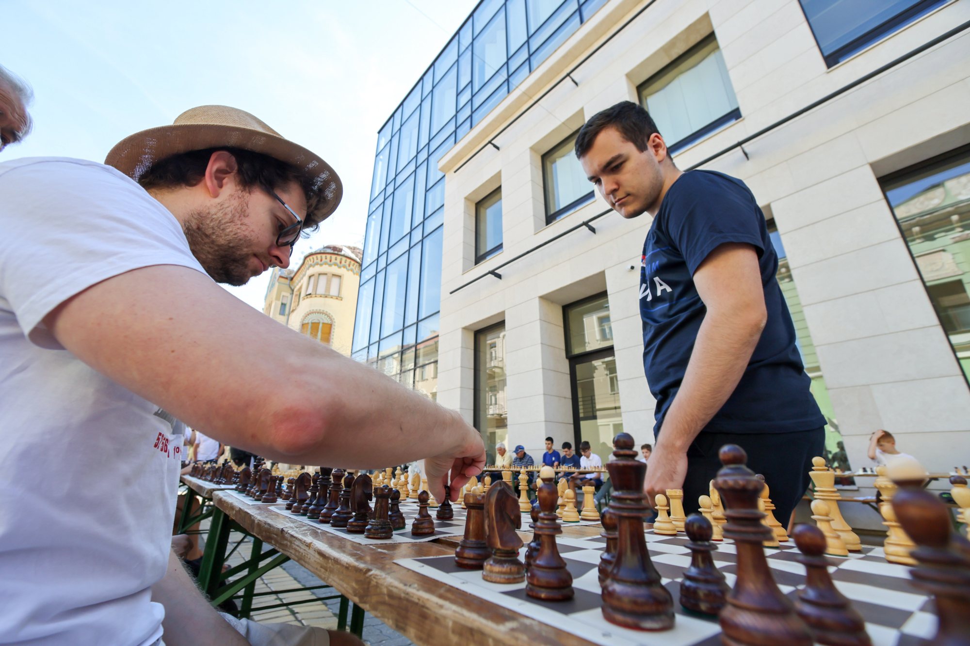 Man in a navy T‑shirt standing at a long row of chessboards on folding tables outdoors; opponent in a white T‑shirt and straw hat reaches to move a piece; modern building façade and a church tower visible behind.
