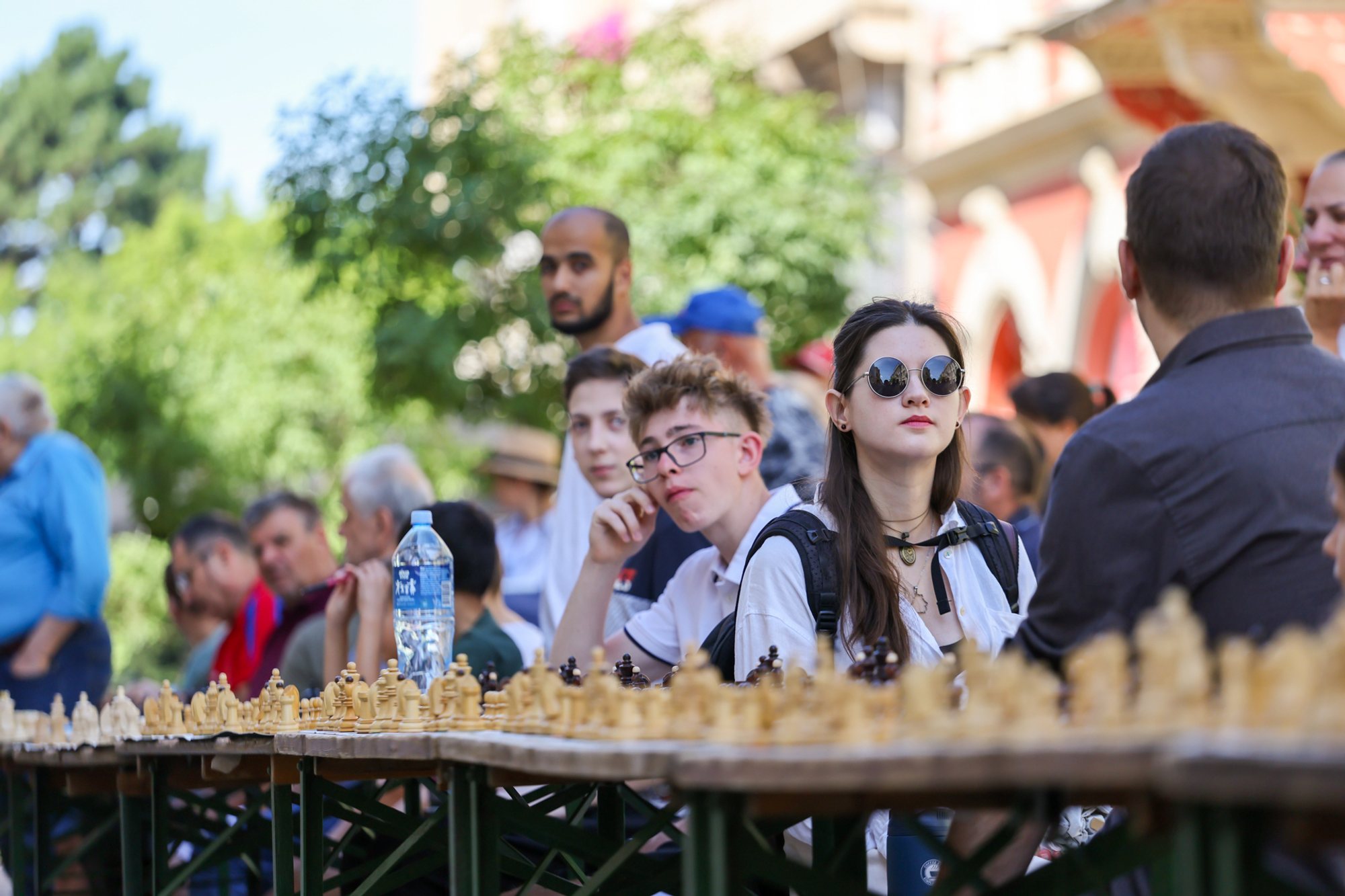 Row of outdoor chessboards on a pedestrian square; three seated players at the nearest table — one looking at the board, one wearing sunglasses — plastic water bottle and score sheets on the table; onlookers and city buildings in soft focus behind.