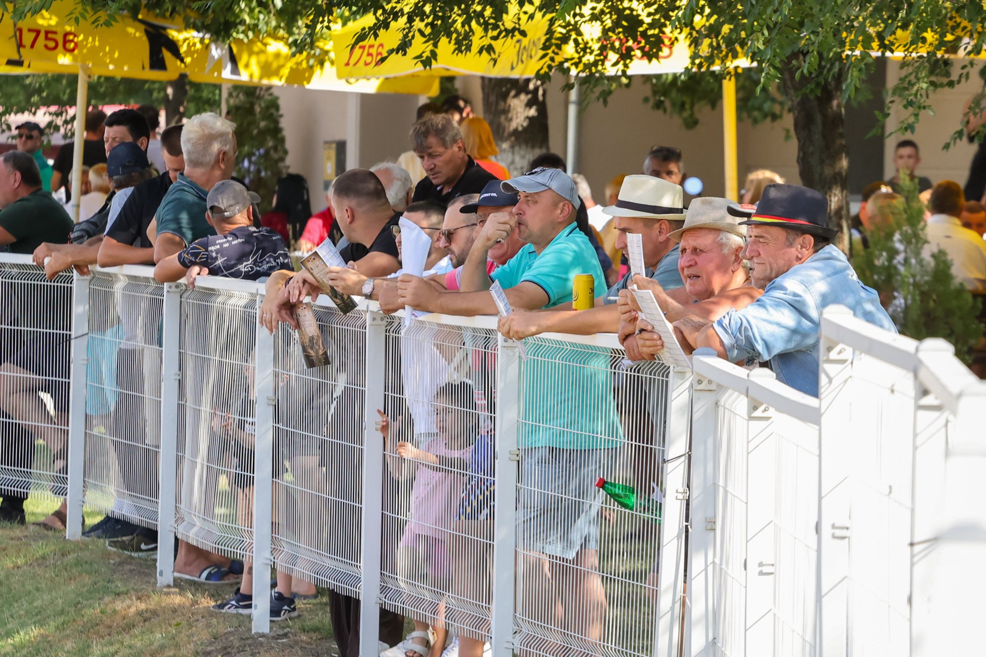 Spectators lined along a white fence at the Subotica hippodrome, shaded by trees and yellow awnings.