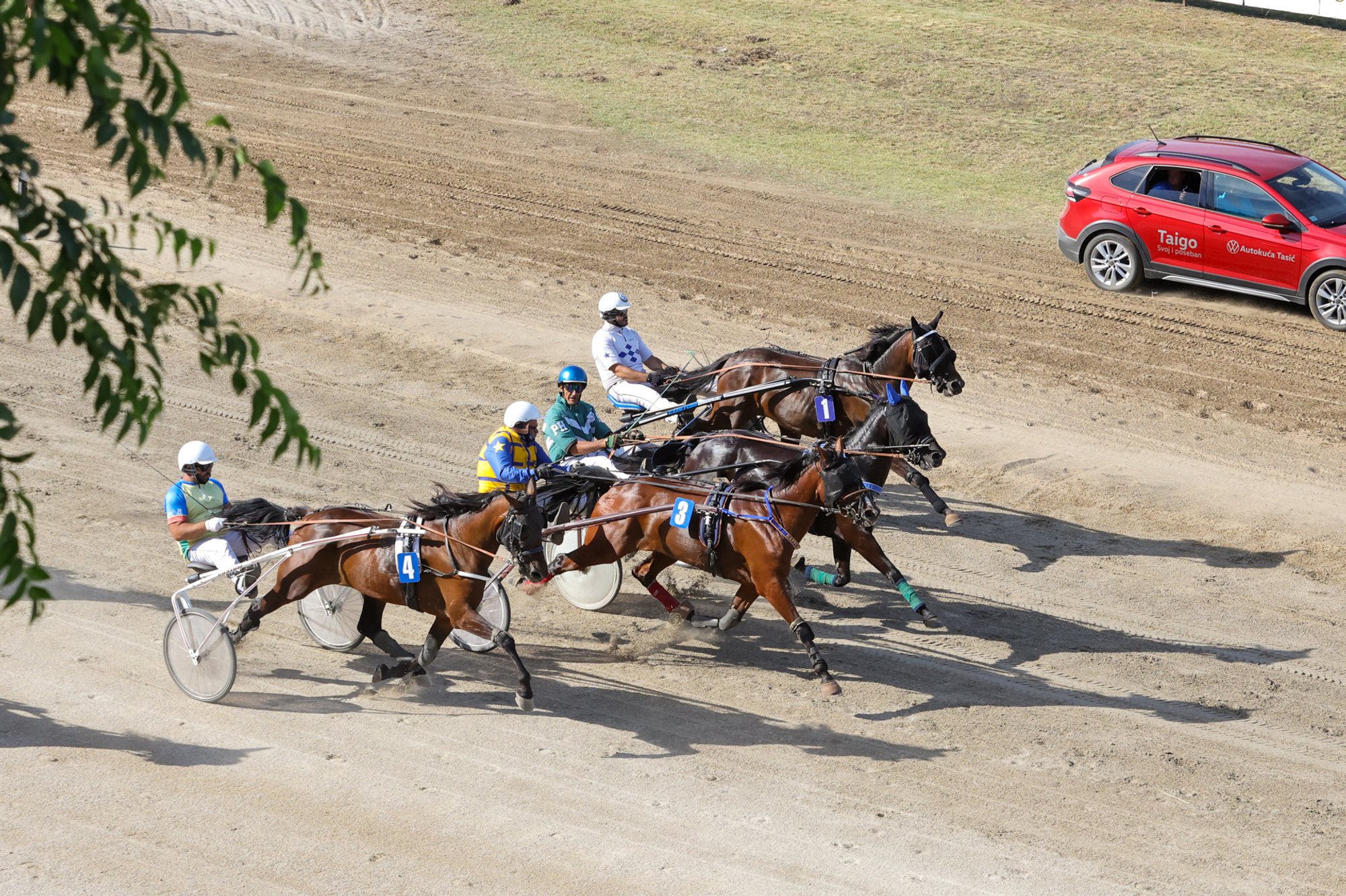 Four trotting horses with people in sulkies racing on a dirt track seen from above; a red car is parked on the infield.