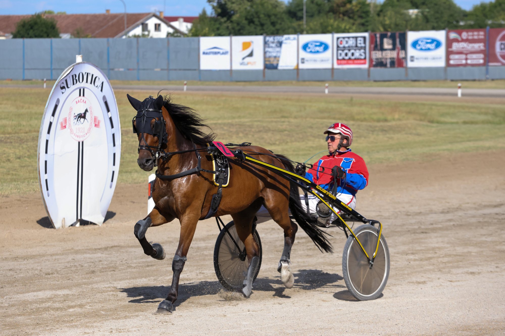 Brown trotting horse in harness pulling a two‑wheeled sulky with a person seated in it on a dirt racetrack; advertising boards and a white starting marker visible behind.