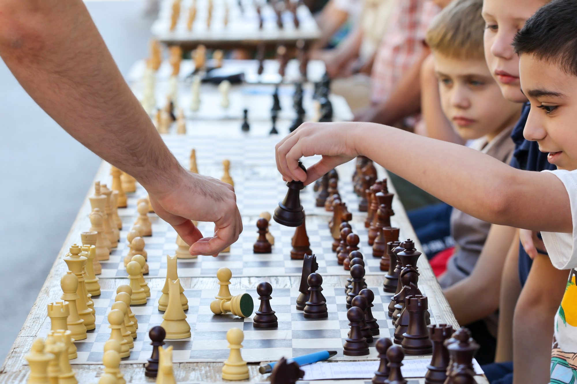 Long row of chessboards on an outdoor table at Subotički Korzo, young participants seated along the table while hands move pieces across multiple boards.