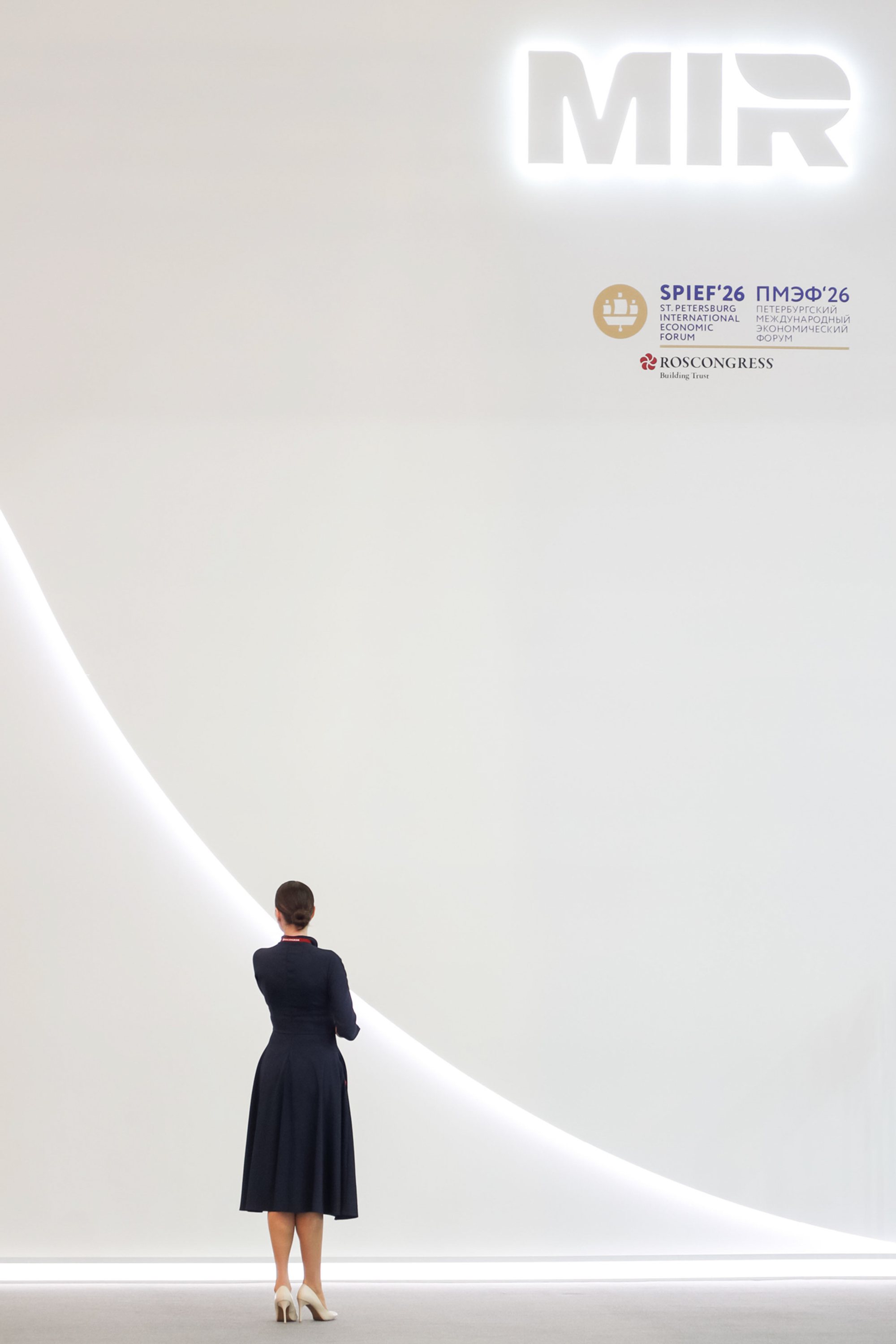 Person in a dark dress standing with hands clasped behind their back, facing a large white exhibition wall with a curved illuminated line and an illuminated 'MIR' sign; small SPIEF and Roscongress logos visible at upper right.