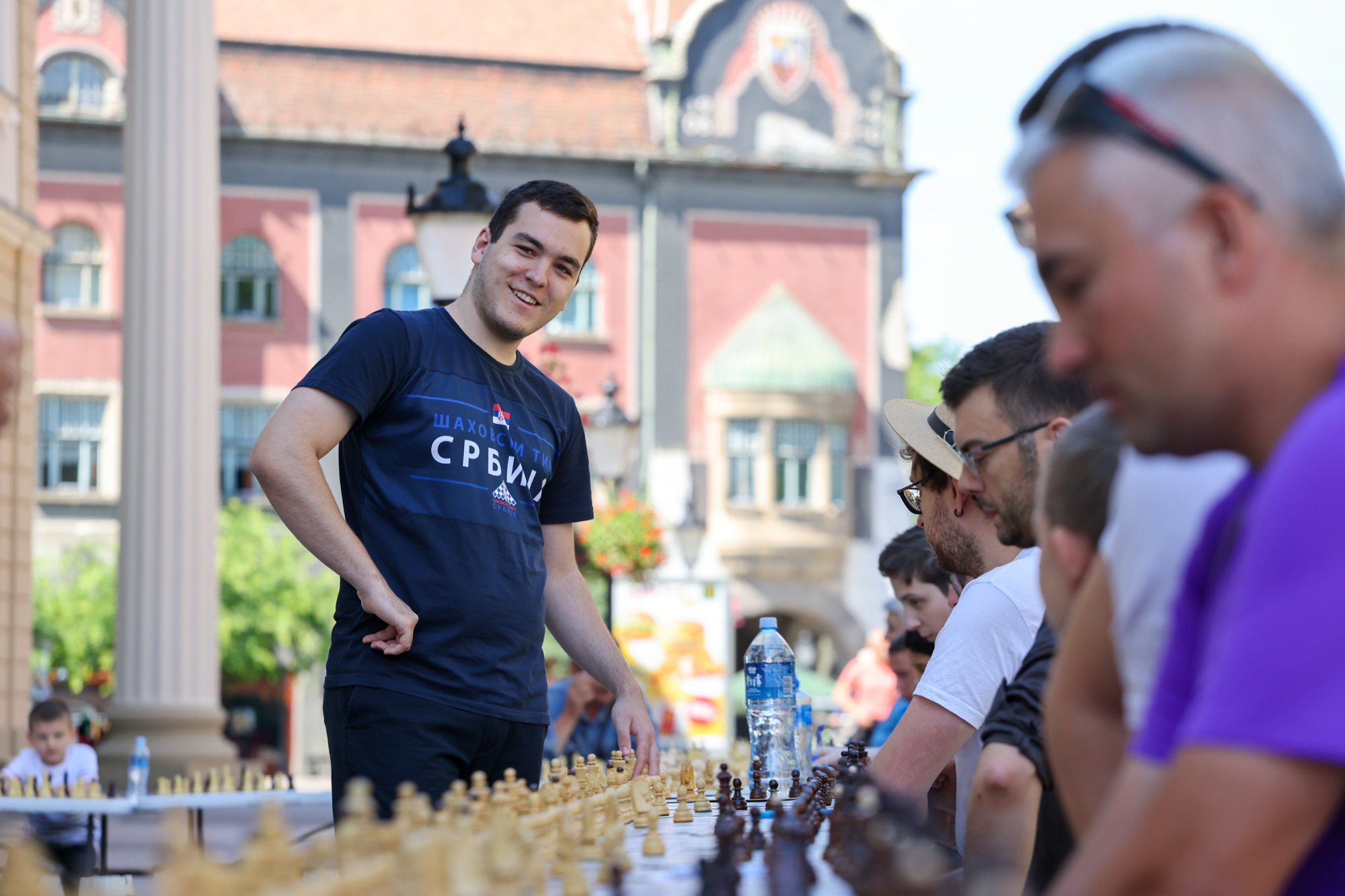 Long row of chessboards on a table along a pedestrian square; a man in a dark T‑shirt with Cyrillic lettering stands beside seated players, historic building façades in the background.