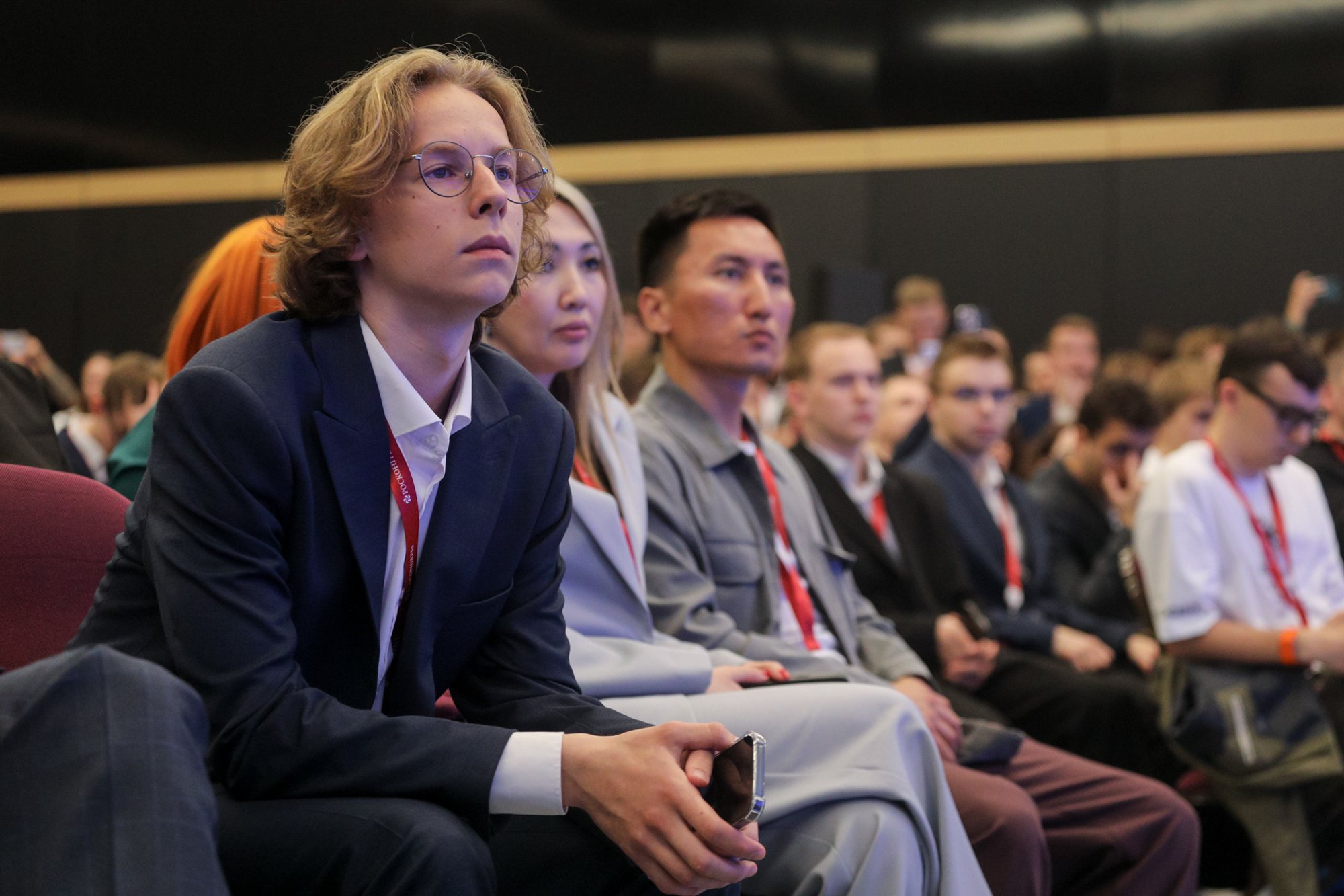 Conference attendees seated in auditorium rows; foreground shows a light‑haired person in a dark blazer holding a smartphone, several people wearing red conference lanyards.