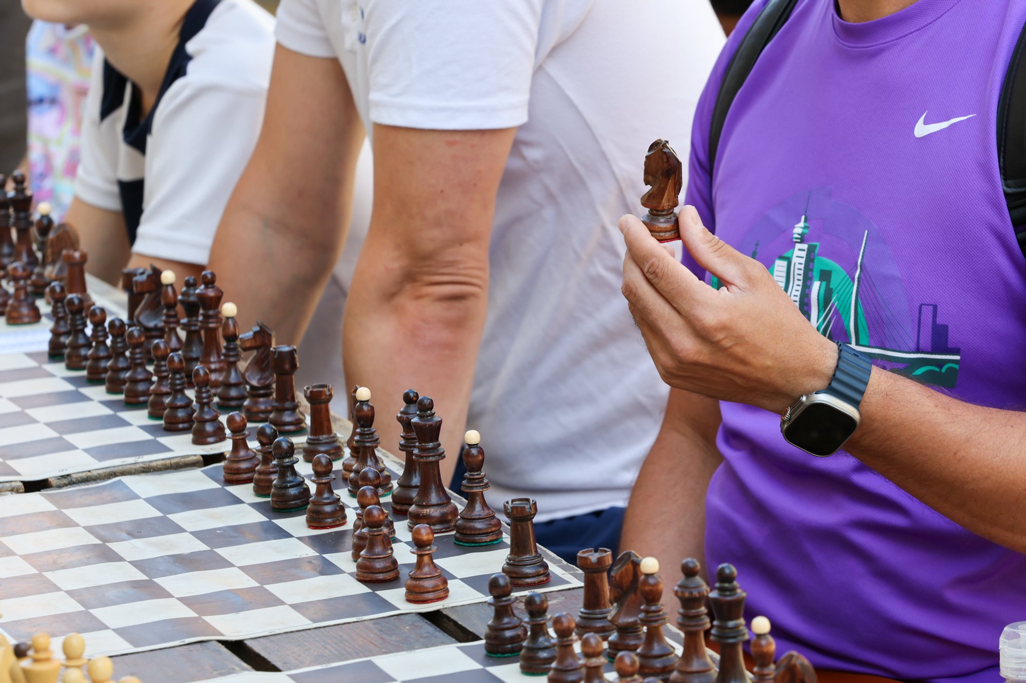 Row of chessboards on a long table with carved wooden pieces; a person in a purple shirt holds a knight.
