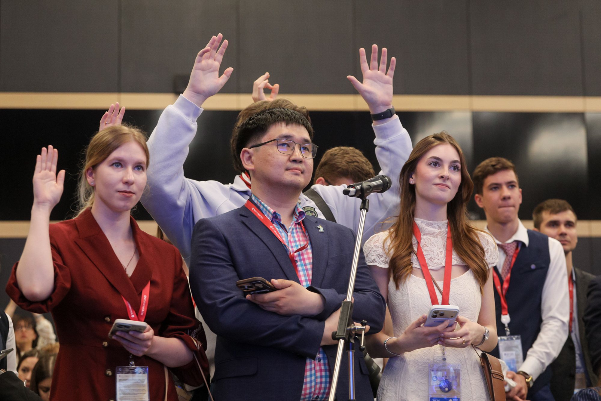 Group of conference attendees standing at a microphone; three people in the foreground wear lanyards — a man holding a smartphone between two women — with several raised hands visible behind them.