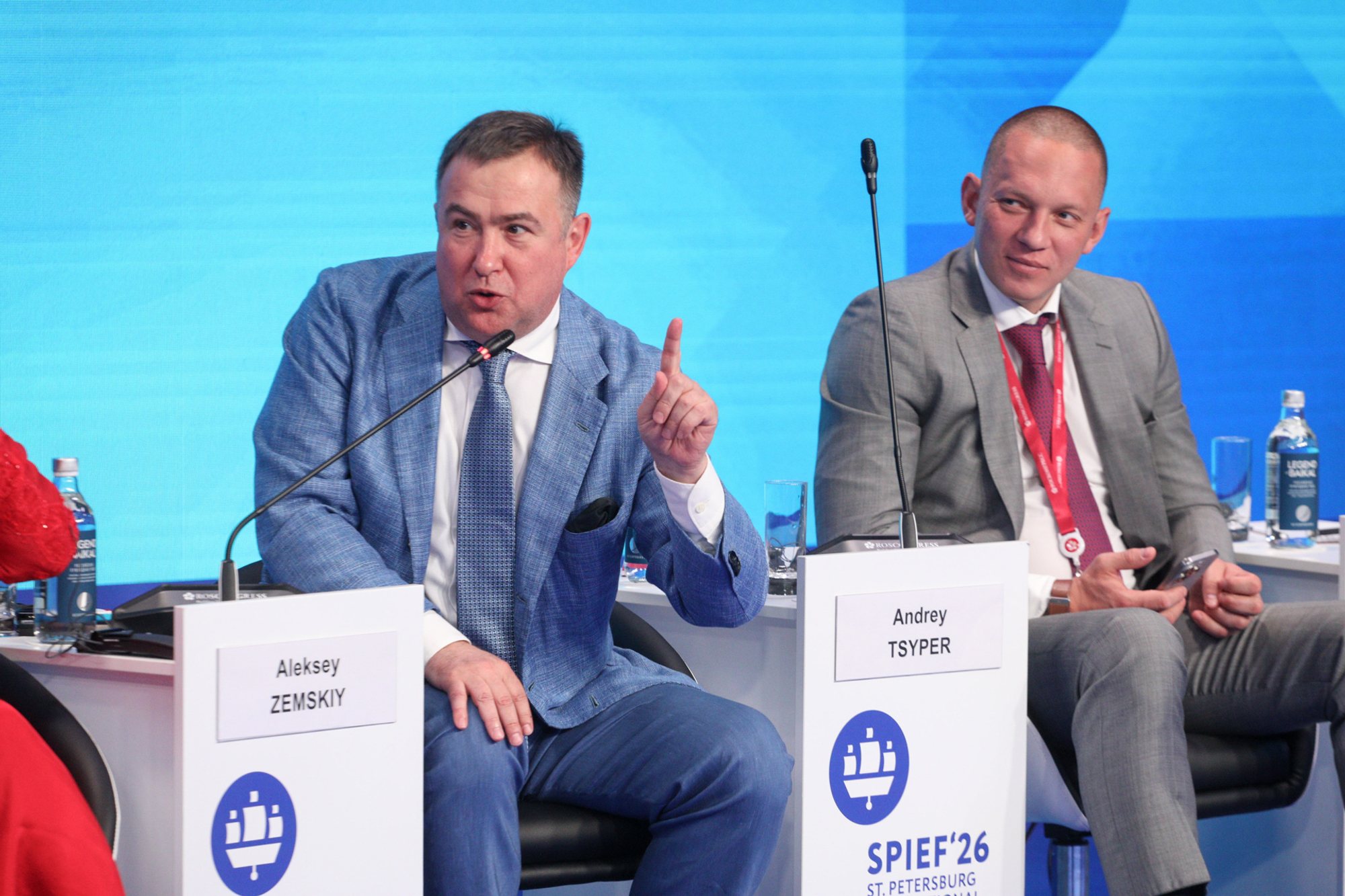 Two suited men seated on a conference panel against a blue stage backdrop, one speaking and gesturing; microphones, bottled water and nameplates on small stands in front of them.