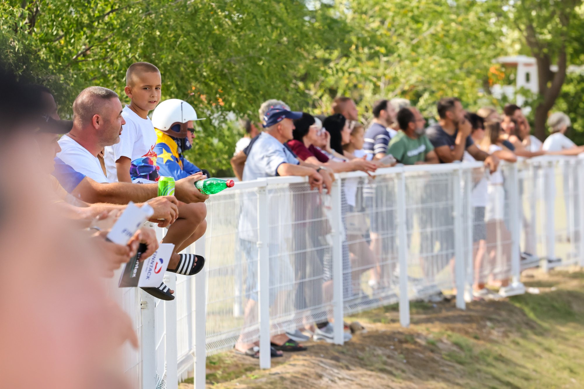 Spectators standing along a white barrier under trees at the edge of Subotica hippodrome, watching trotting races.