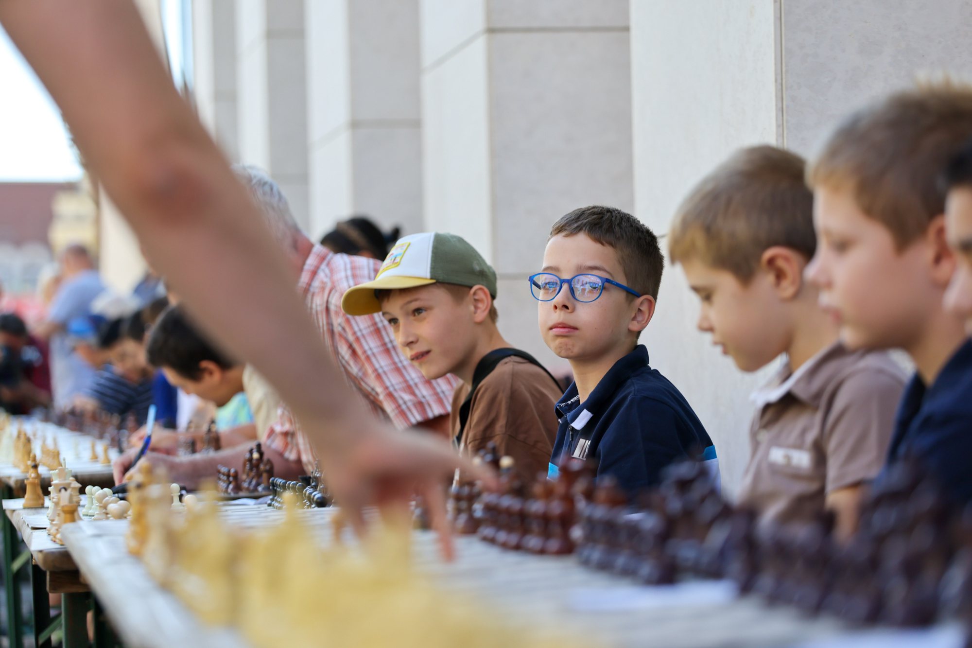 Long line of chessboards on a pedestrian square under a colonnade; children seated at the boards while a standing person's arm reaches across a board to make a move.