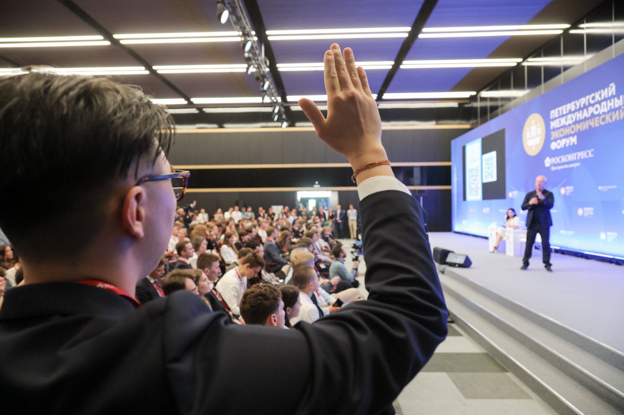 Conference auditorium at ExpoForum: a person in the foreground raises a hand toward a speaker on stage; tiered seating filled with attendees.