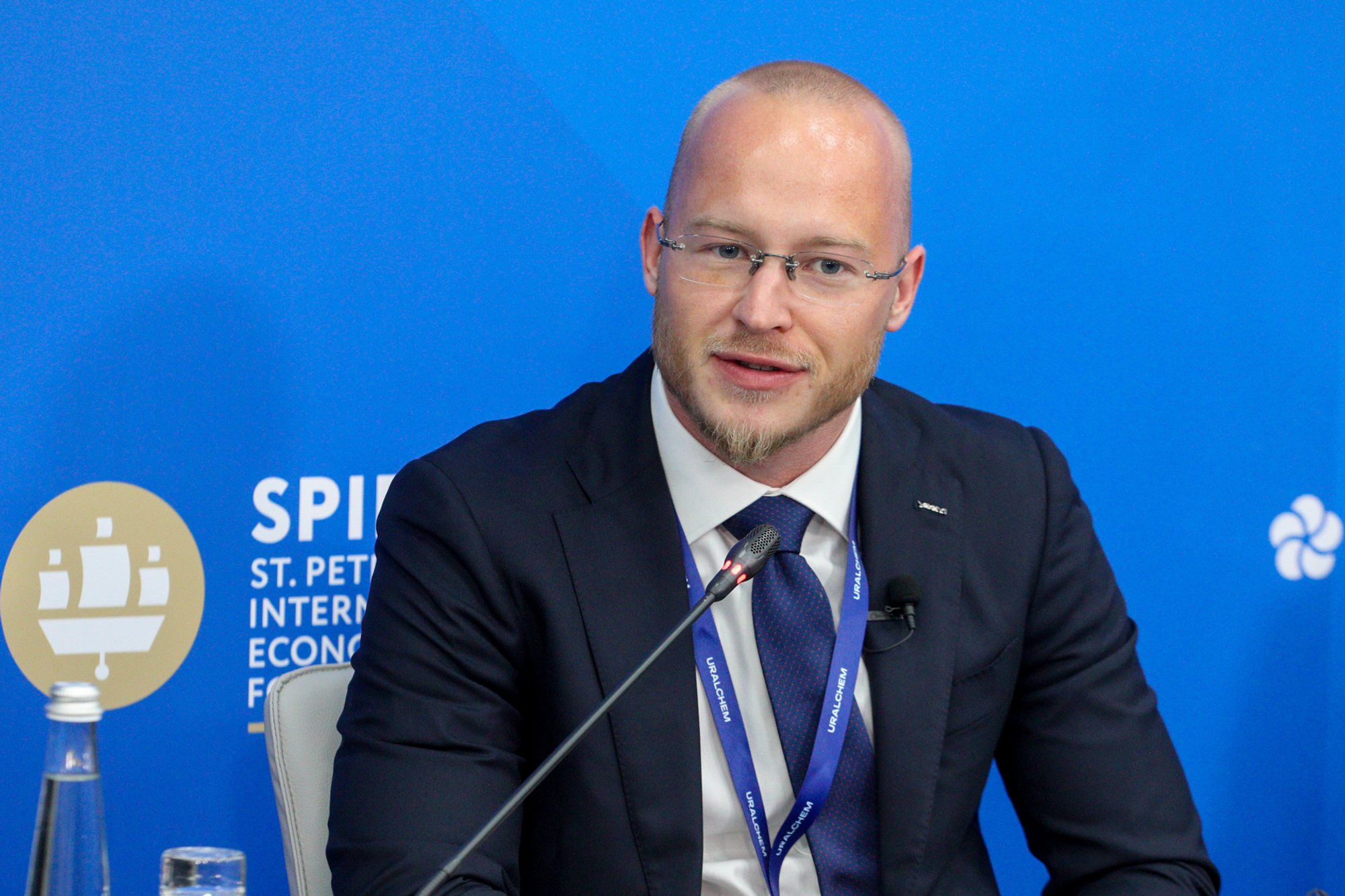 Person in a dark suit and glasses speaking into a tabletop microphone against a blue backdrop with 'SPIEF' branding; conference lanyard and a water glass on the table.