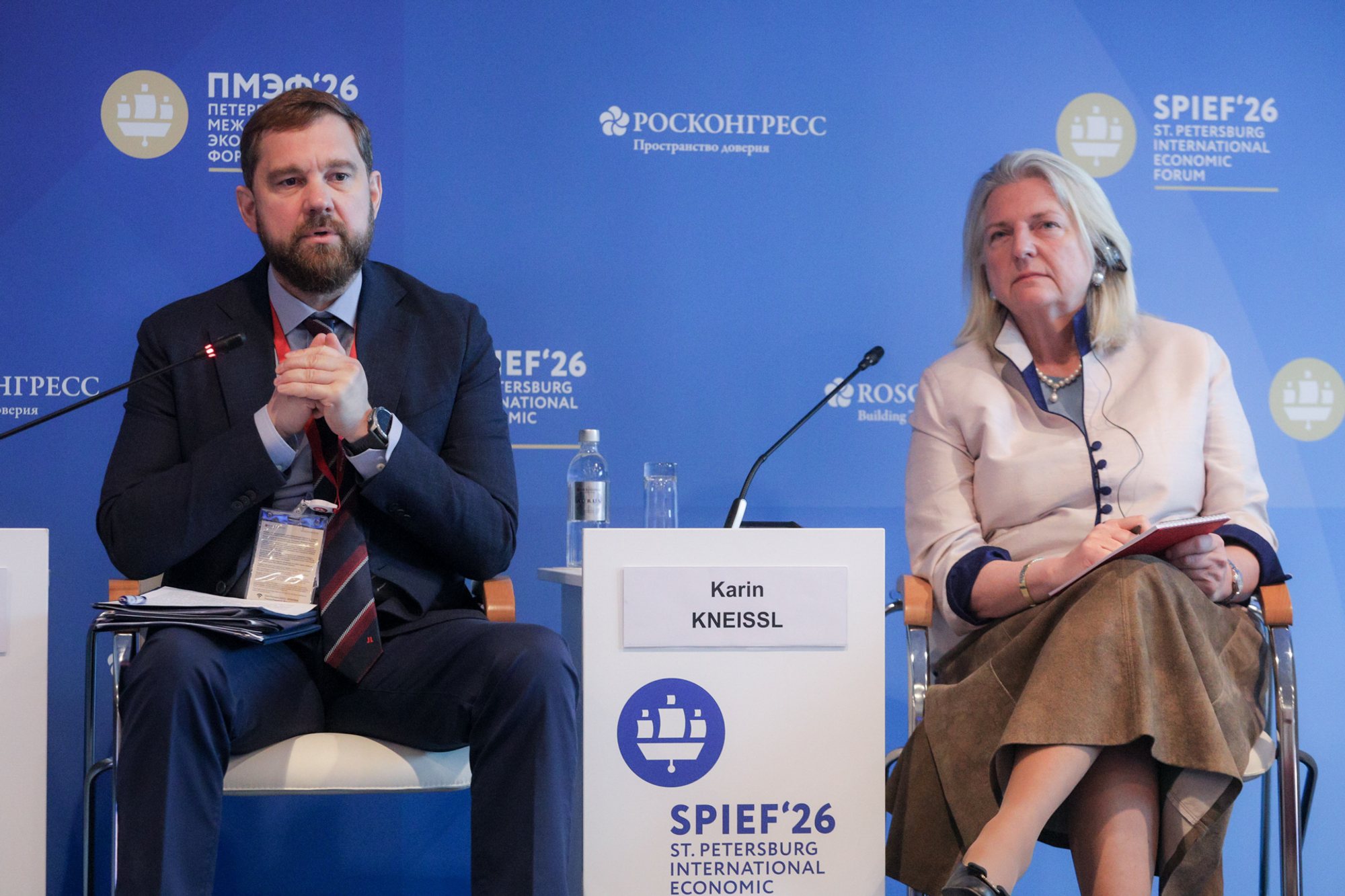 Two seated panelists on a blue stage backdrop with event and organizer logos, microphones on stands, a small branded cube table and a visible nameplate (not transcribed).