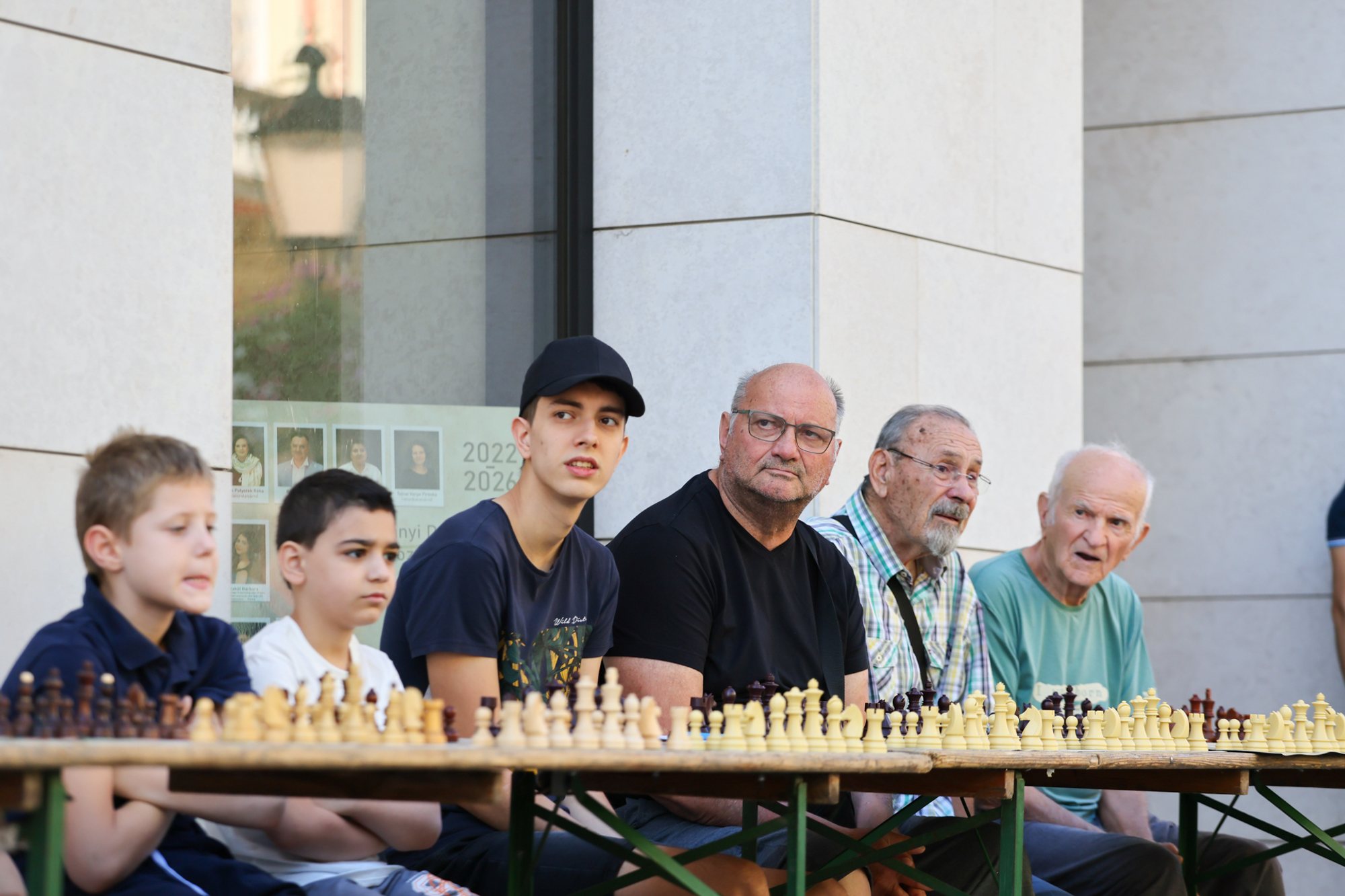 Six people seated along a long table with multiple chessboards and pieces set up outdoors in front of a pale building.