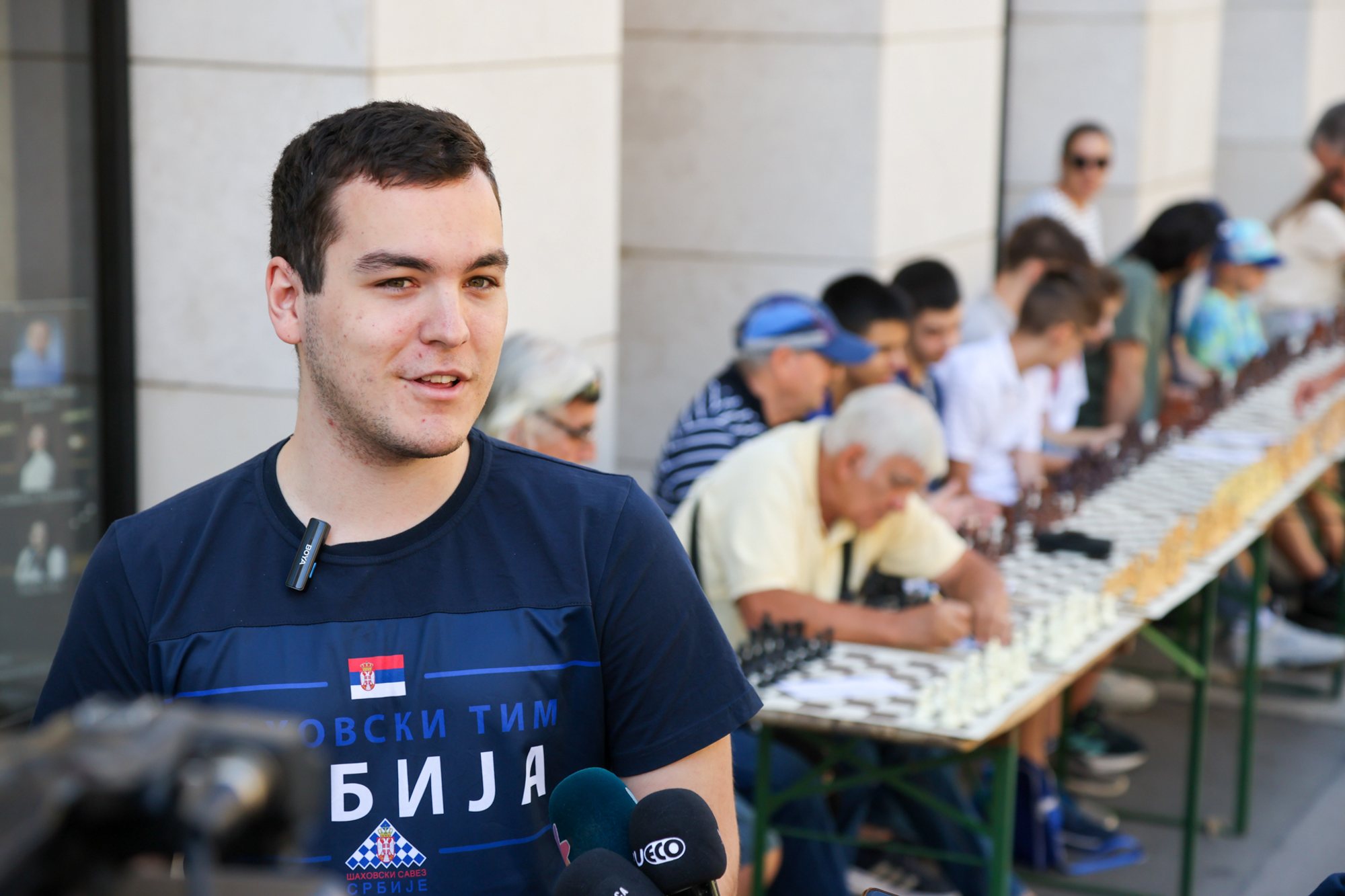 Man speaking into microphones with a long row of seated chess players and boards along a pedestrian square in the background.