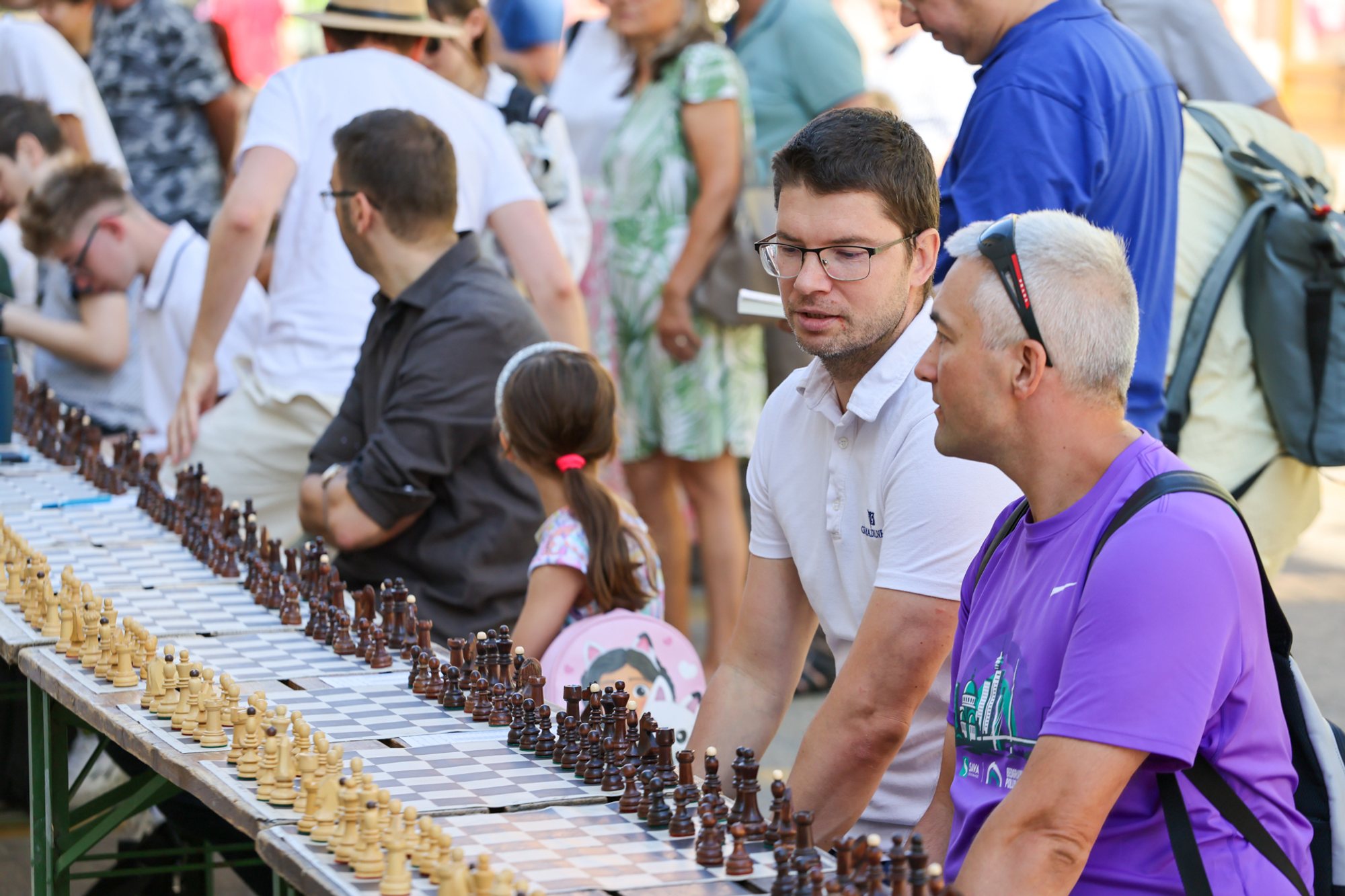 Long row of chessboards on folding tables along a busy pedestrian square; players and spectators sit and stand beside the boards.