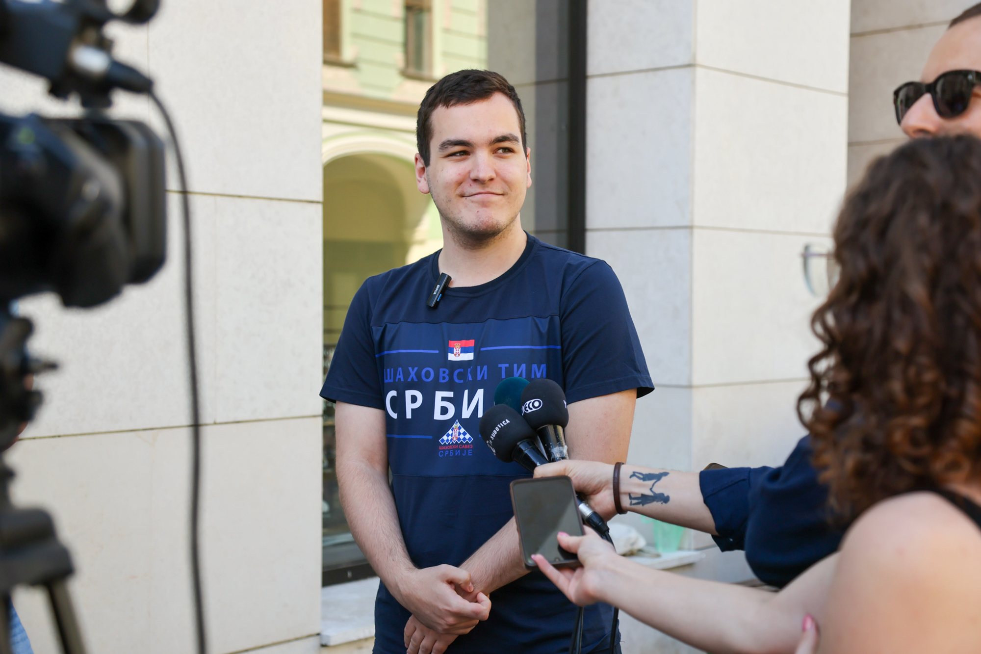 Person in a dark T‑shirt with Cyrillic 'СРБИ' stands beside a column while reporters hold microphones and a video camera; scene from a chess simultaneous on Subotički Korzo.
