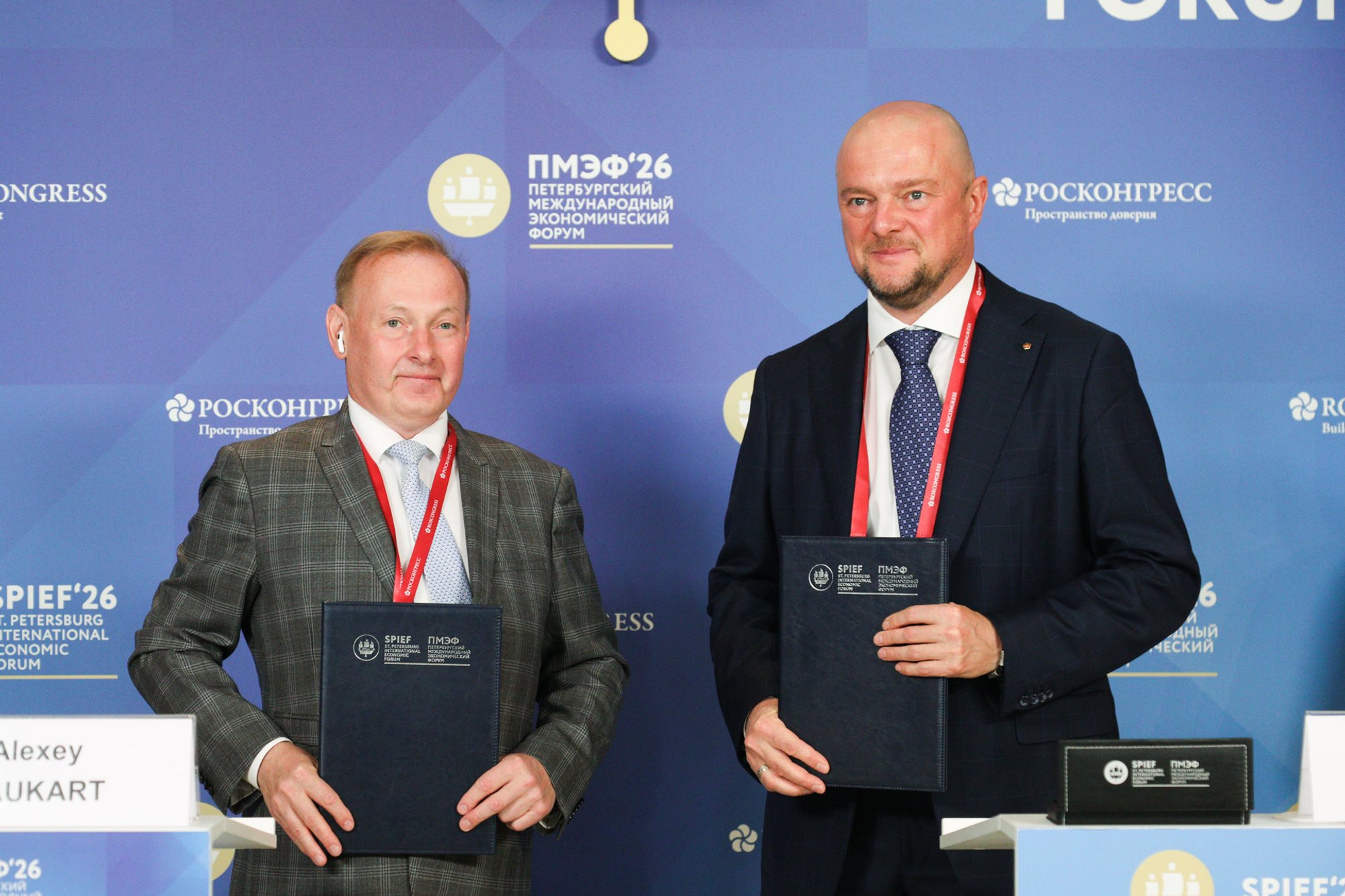 Two men standing side by side holding dark folders in front of a blue backdrop with SPIEF and Roscongress logos.