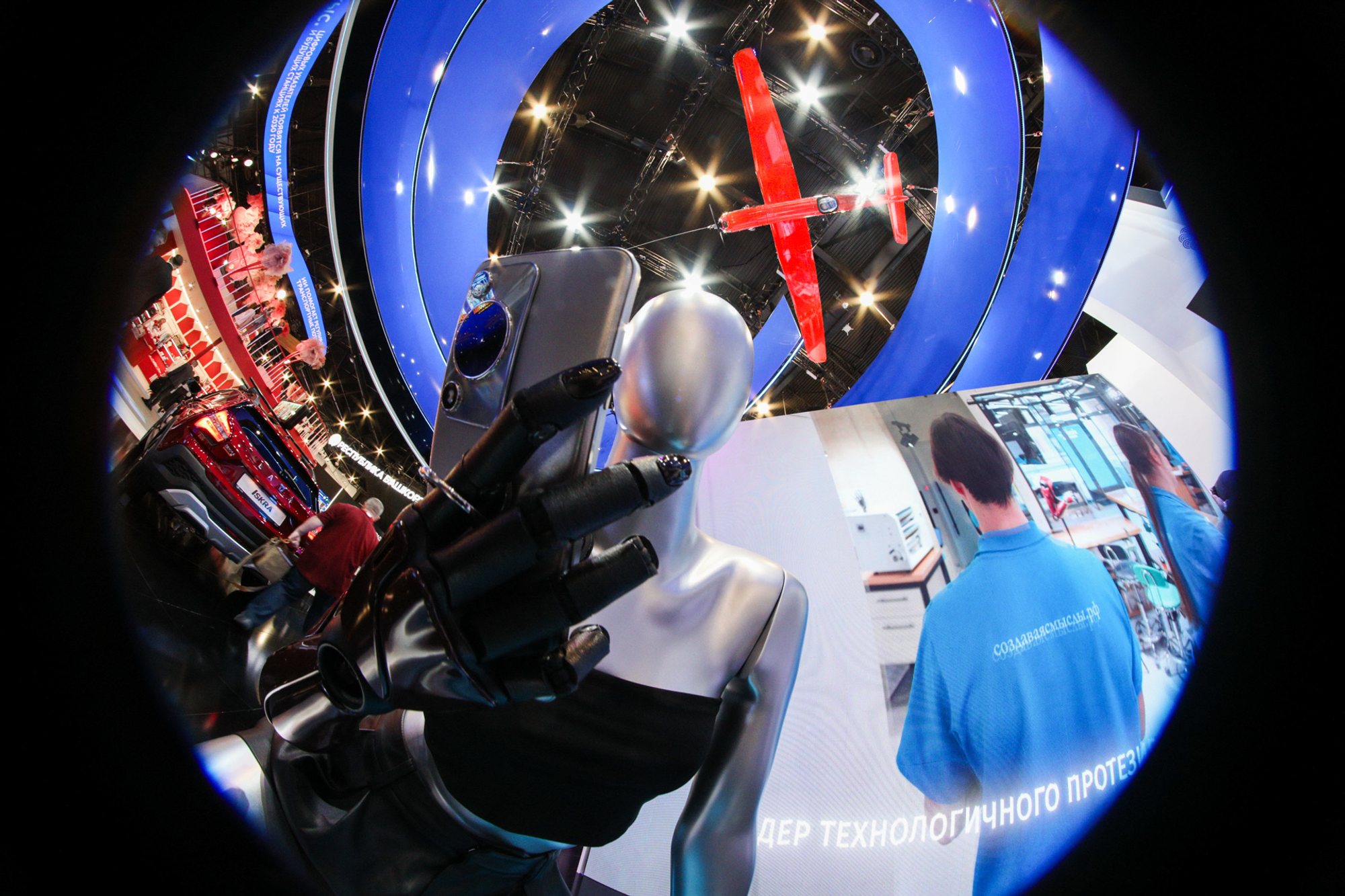 Fisheye view of a white humanoid robot extending its hand toward the camera at a blue-curved exhibition stand, with a person in a blue jacket viewing digital displays and spotlights overhead.