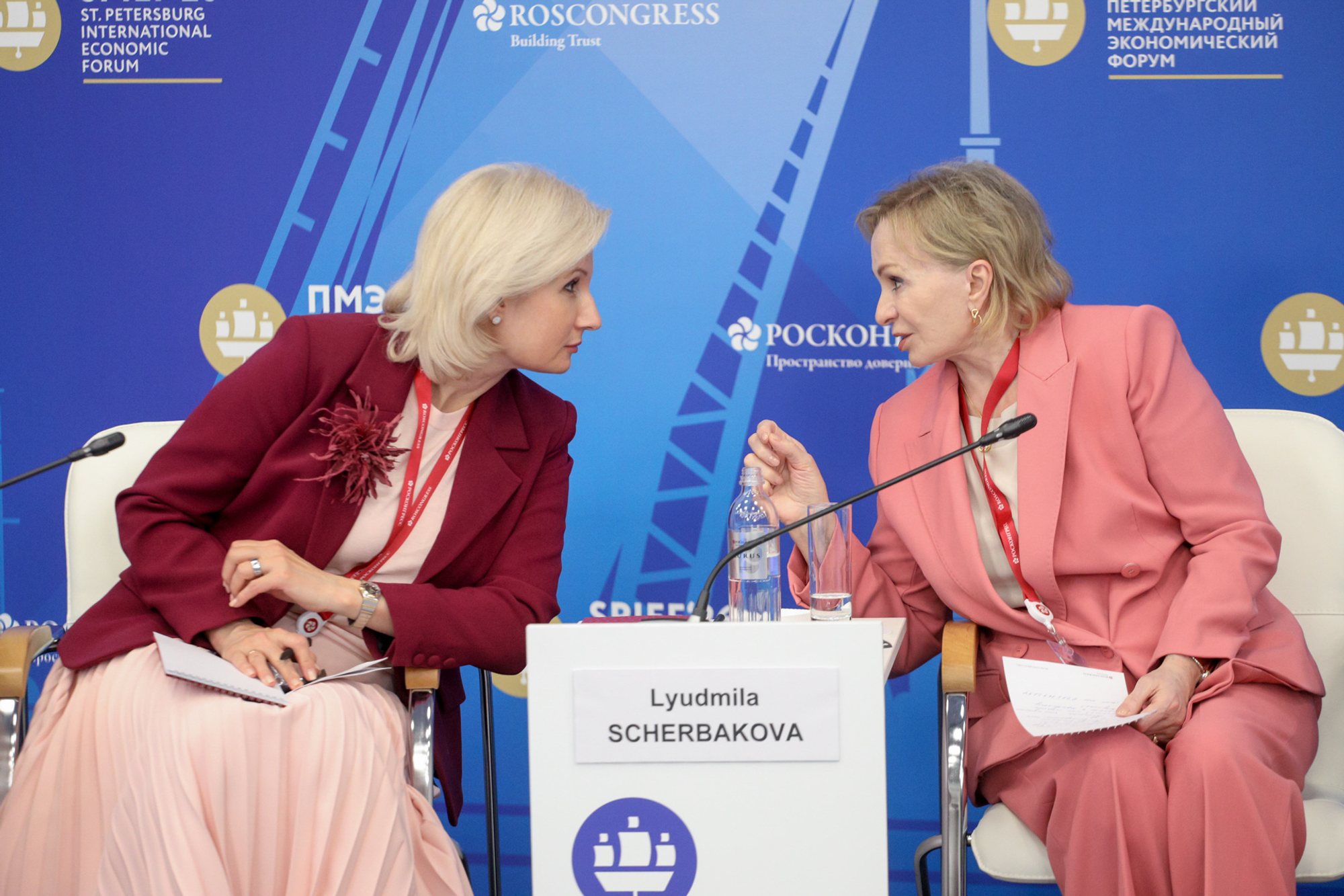 Two women seated at a panel table, turned toward each other and speaking into tabletop microphones; a white placard with printed text and a blue conference backdrop with logos are visible behind them.