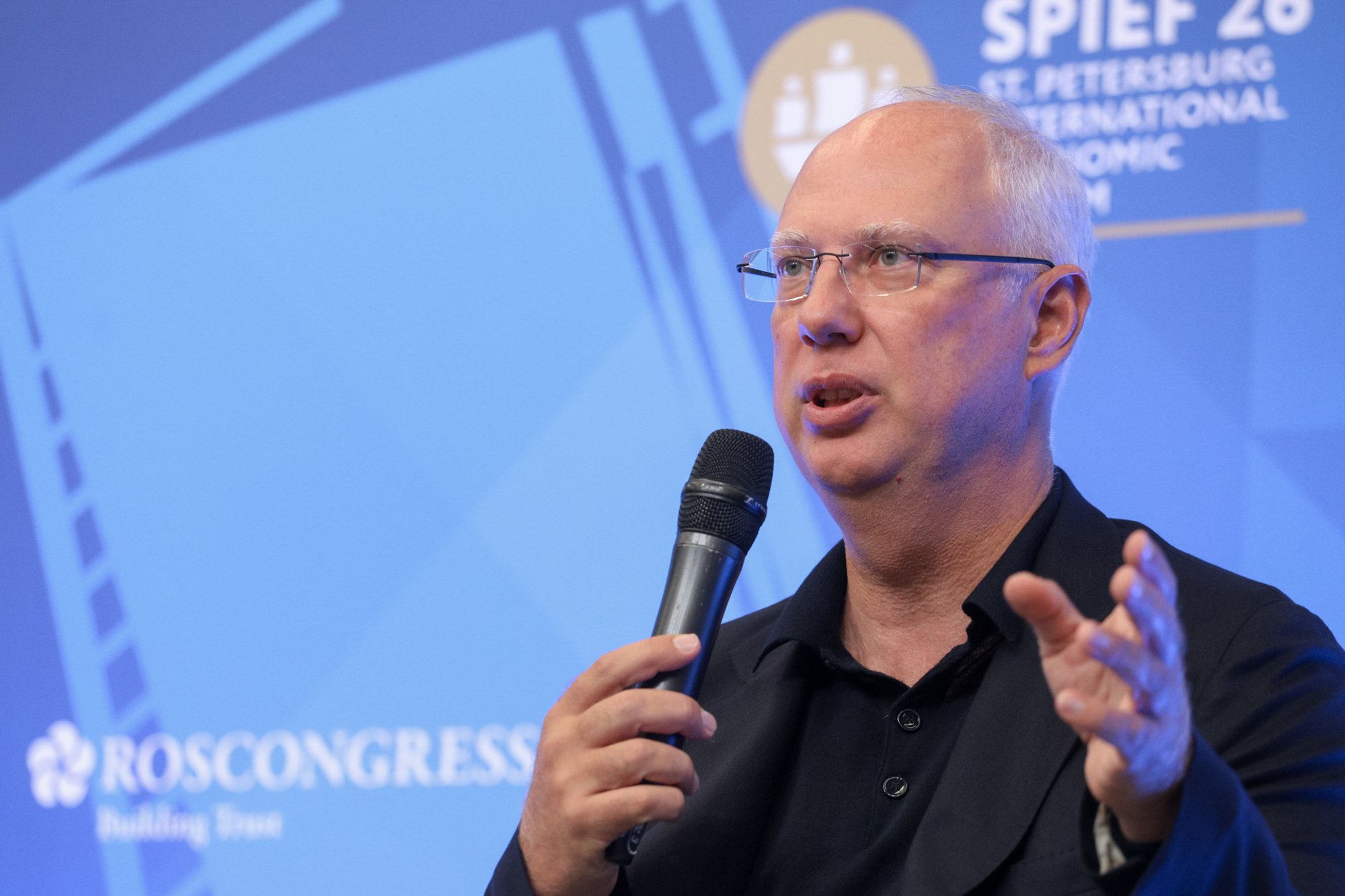 Man wearing glasses and a dark jacket holds a handheld microphone and gestures in front of a blue stage backdrop showing SPIEF signage and the Roscongress logo.