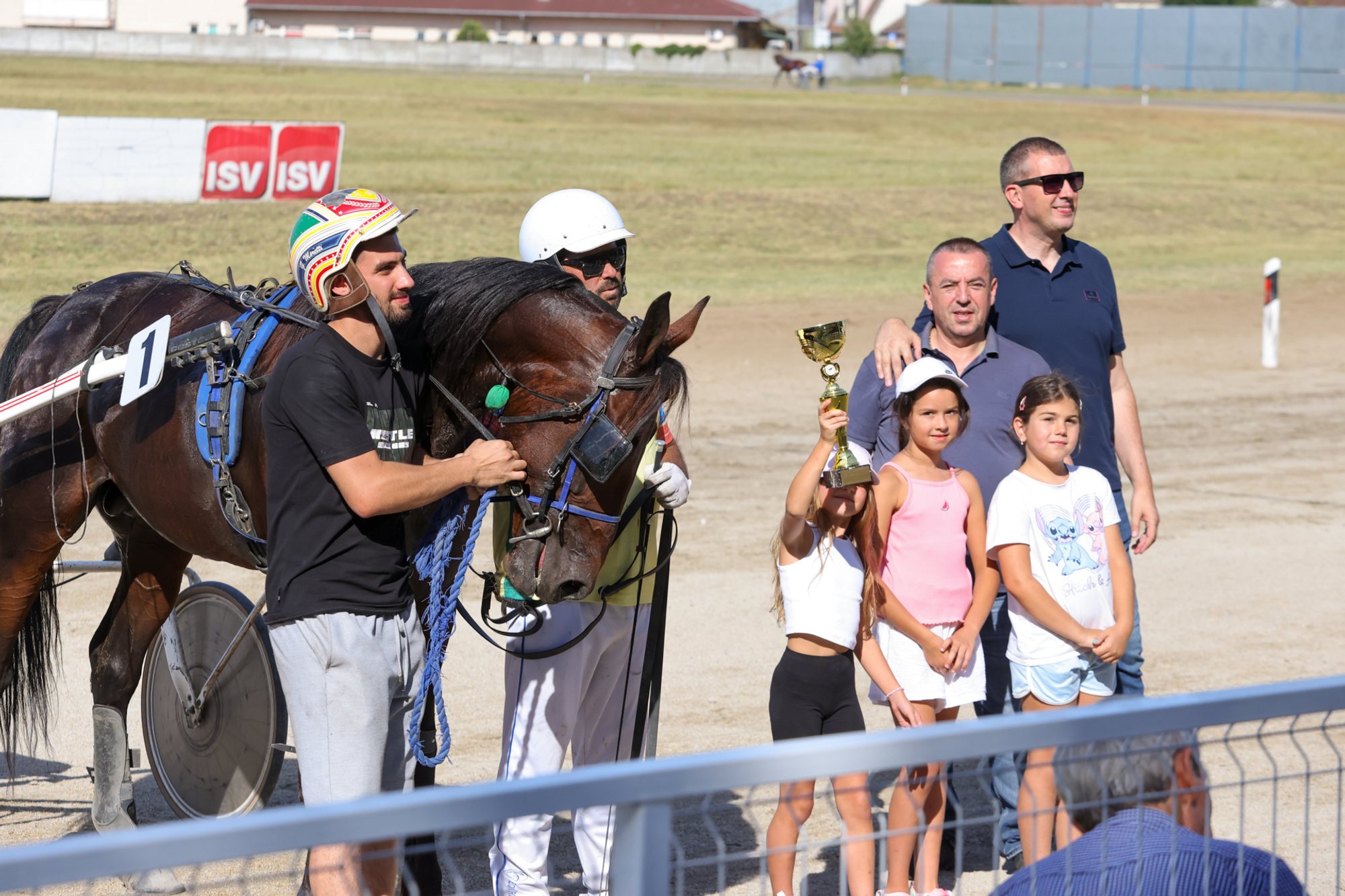 Dark trotting horse wearing saddlecloth number 10 at a metal railing on a dirt racetrack; two adults hold the horse’s bridle while four people — three children holding small trophies — stand beside the railing, with the track and sponsor boards visible in the background.